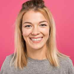 A young woman with straight blonde hair, wearing a grey shirt, smiles at the camera against a solid pink background.