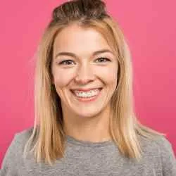 A young woman with straight blonde hair, wearing a grey shirt, smiles at the camera against a solid pink background.