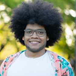 Smiling person with glasses and curly hair, wearing a white shirt and a floral jacket, standing outdoors with green trees in the background.