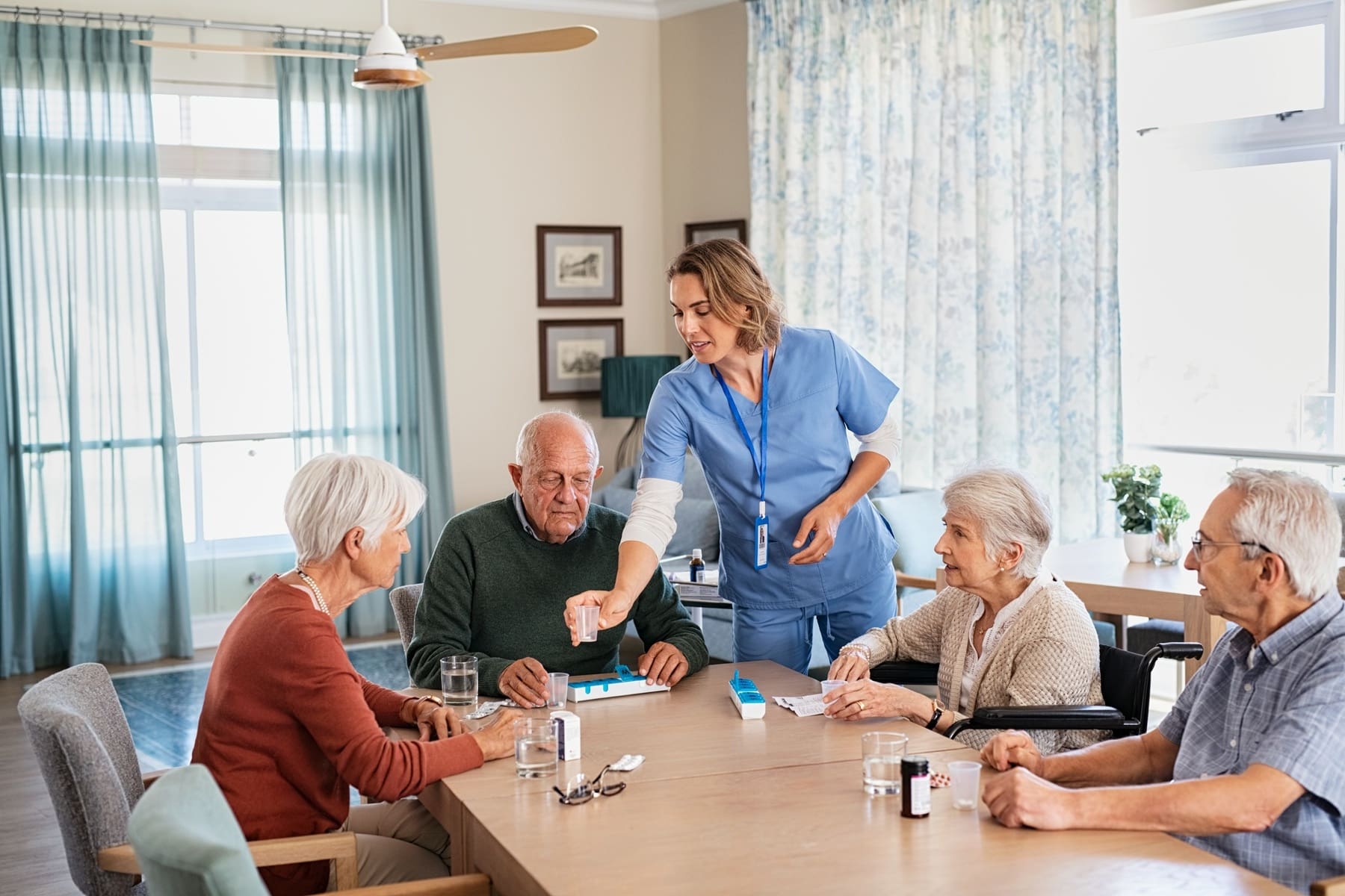 A caregiver in blue scrubs assists four elderly people with medication at a table in a bright, well-lit room.