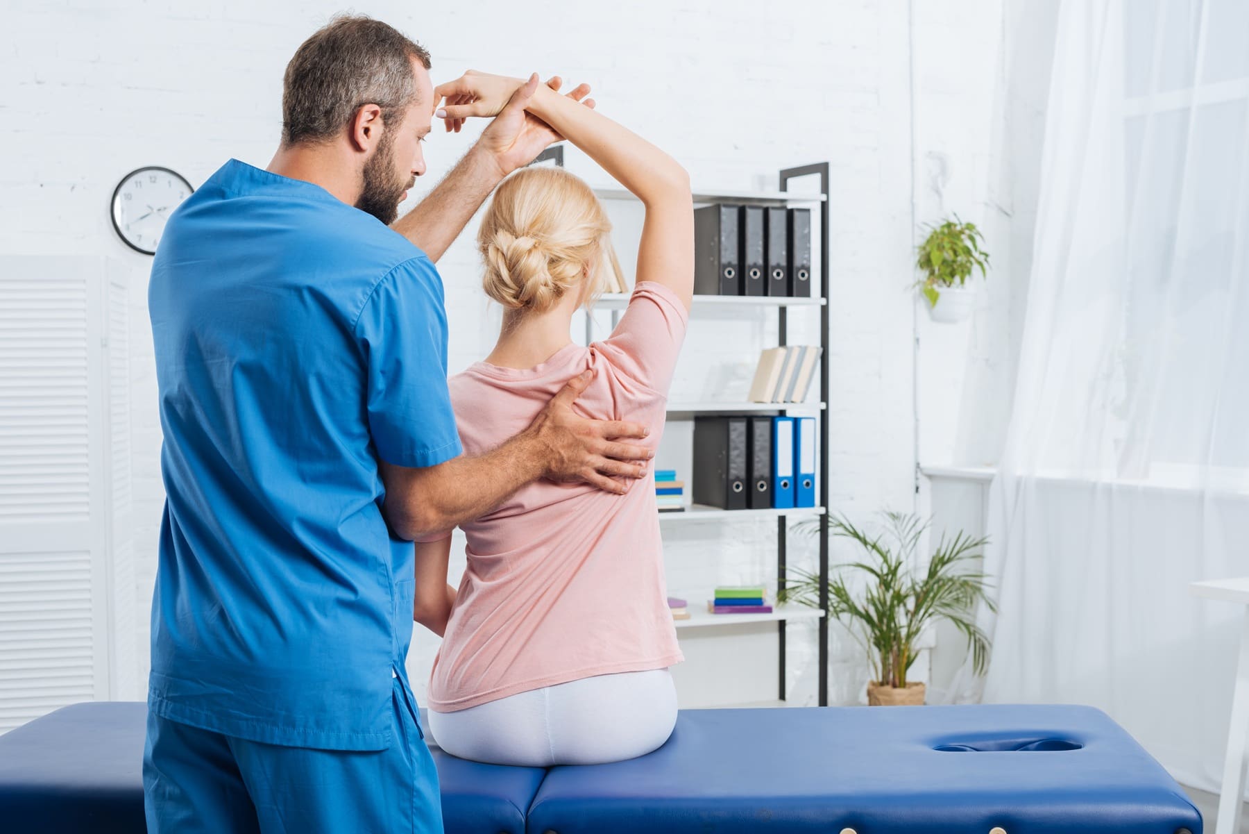 A healthcare professional in blue scrubs assists a seated woman with a shoulder and arm mobility exercise in a medical office.