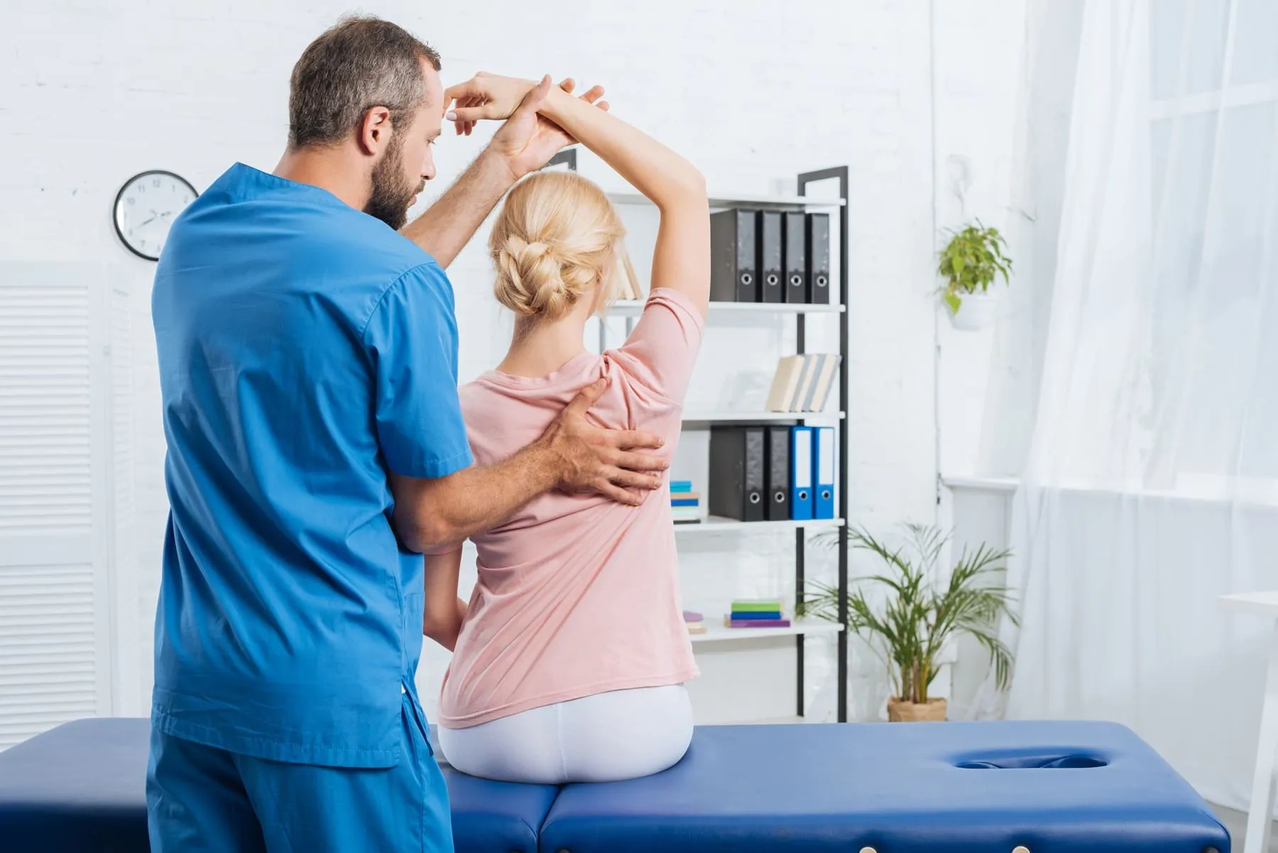 A healthcare professional in blue scrubs assists a seated woman with a shoulder and arm mobility exercise in a medical office.