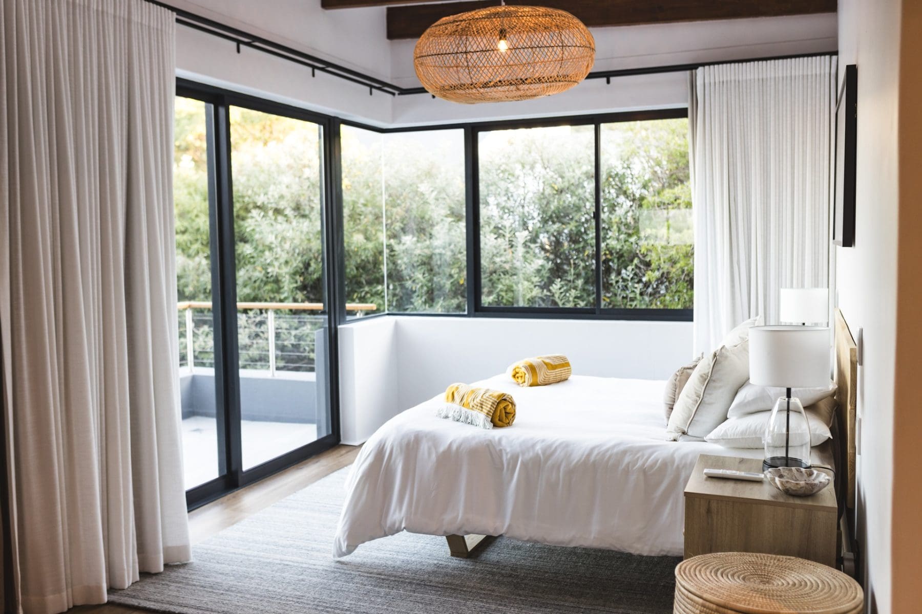 Modern bedroom with large windows, a double bed with white linens and yellow towels, a wooden nightstand, woven ceiling light, and beige curtains. Trees are visible outside.