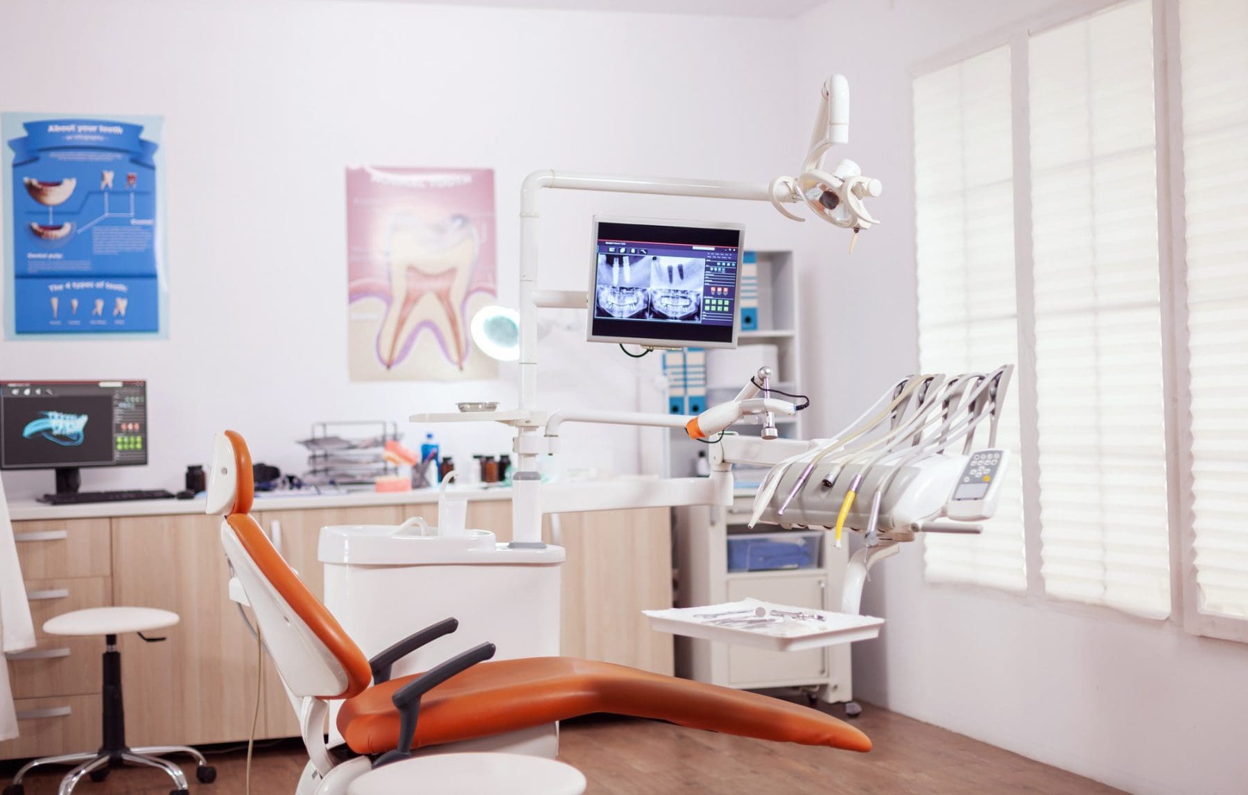 Modern dental office with an orange dental chair, dental equipment, X-ray images on monitors, and posters on the walls. Natural light enters through large window blinds.