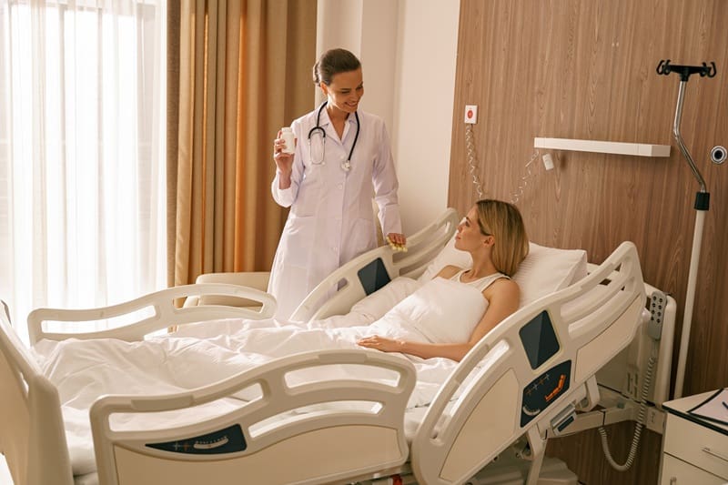 A doctor stands beside a hospital bed, holding medication and speaking to a patient who is lying in bed in a hospital room.