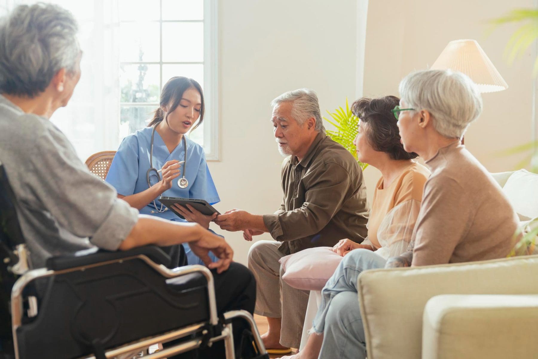 A nurse holding a tablet speaks with four elderly people, one in a wheelchair, in a brightly lit living room.
