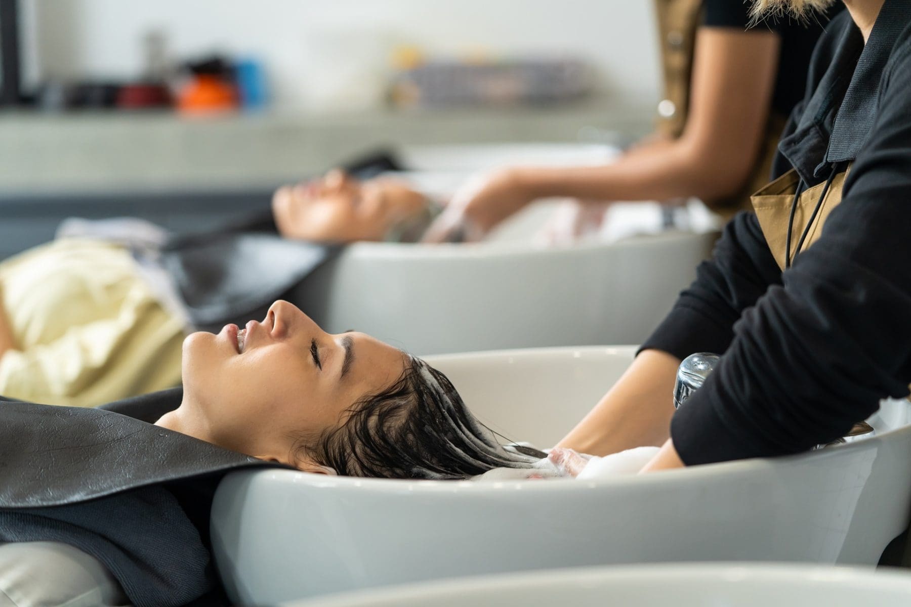 Two people are lying back at salon sinks with their hair being washed by stylists.