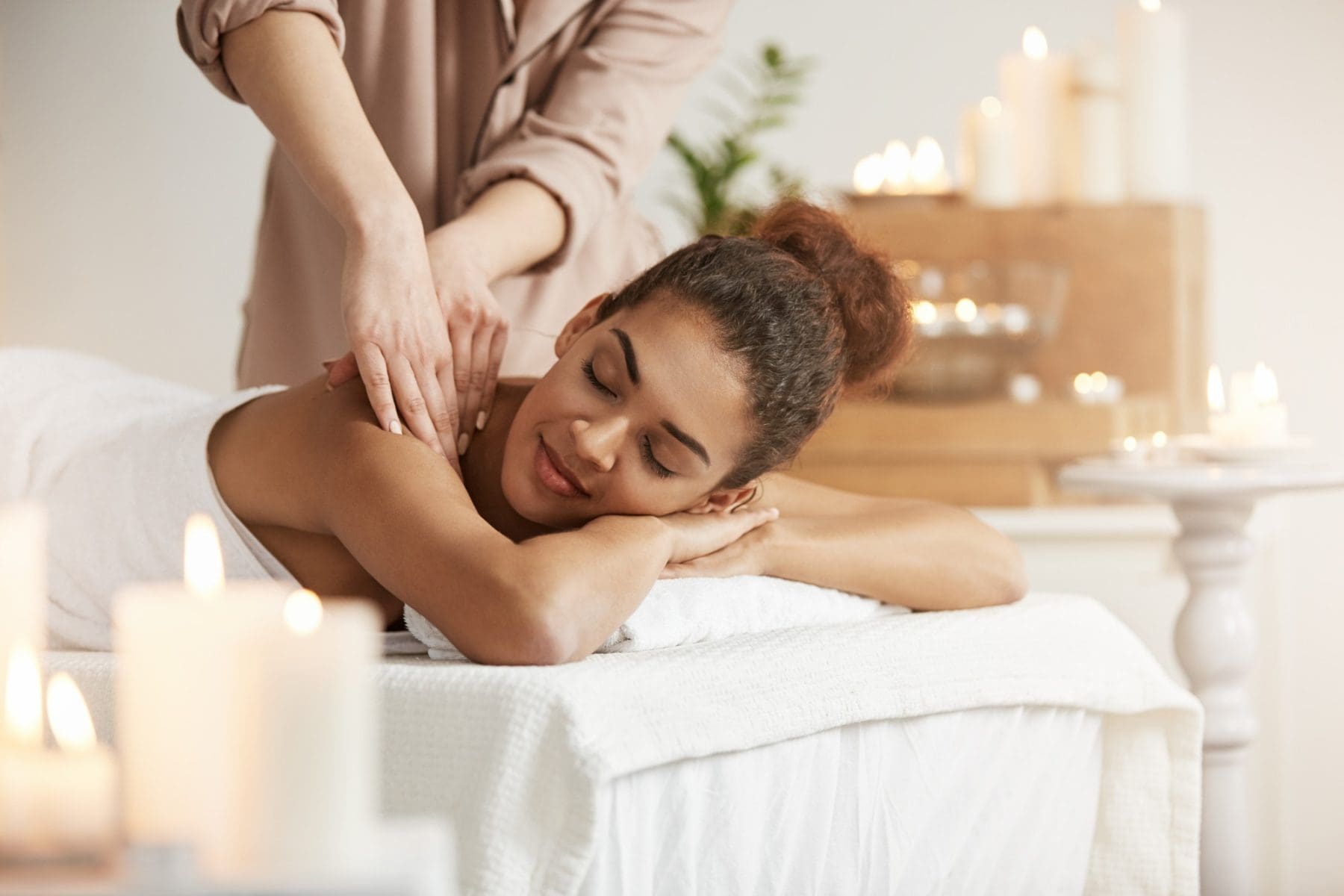 A woman receives a shoulder massage while lying on a massage table, surrounded by lit candles in a calm, softly lit room.
