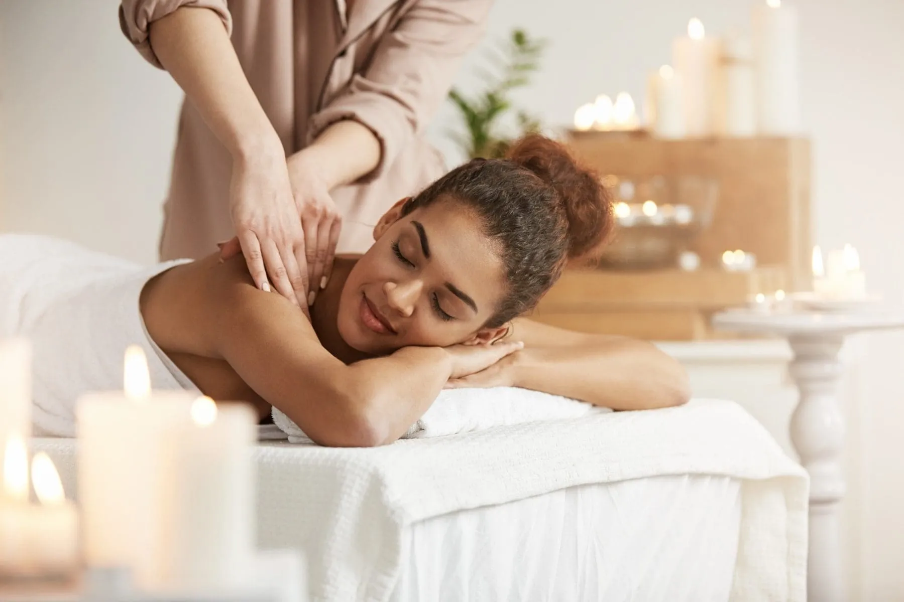 A woman receives a shoulder massage while lying on a massage table, surrounded by lit candles in a calm, softly lit room.