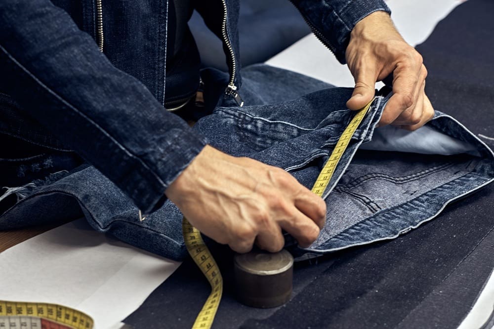 Close-up of a person measuring denim fabric with a yellow measuring tape, wearing a denim jacket, with pattern materials on the table.