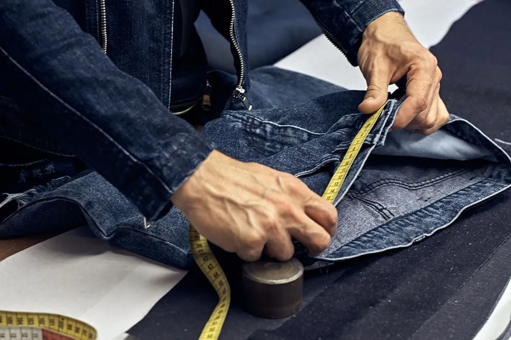 Close-up of a person measuring denim fabric with a yellow measuring tape, wearing a denim jacket, with pattern materials on the table.